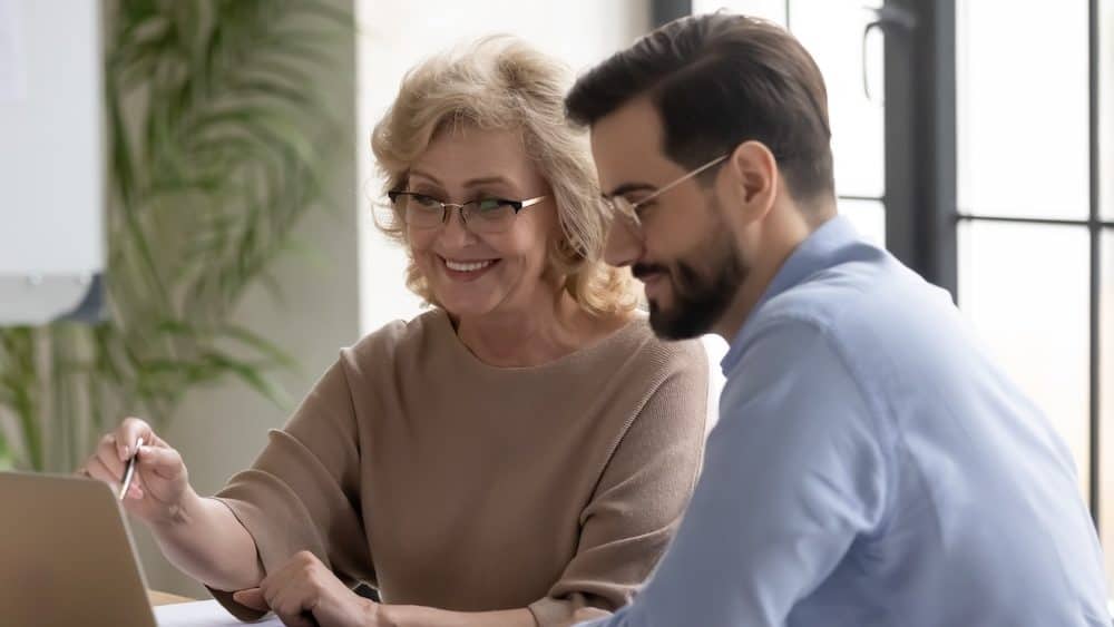 Smiling mature middle aged businesswoman mentor coach teaching intern, pointing at laptop screen, helping with corporate software to new employee, giving instructions, colleagues working together